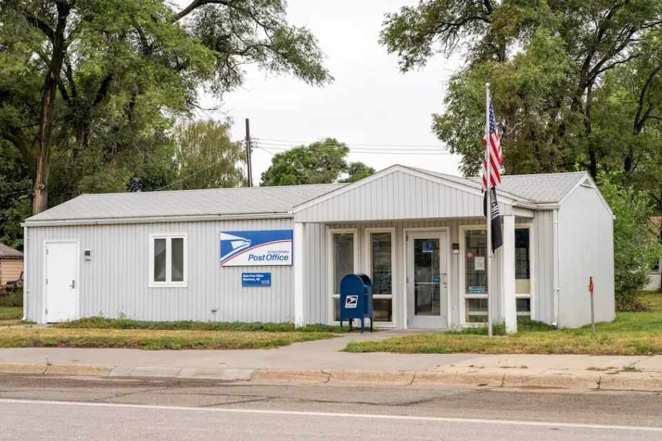 Small rural USPS post office in manufactured one-story grey building with American flag in front.